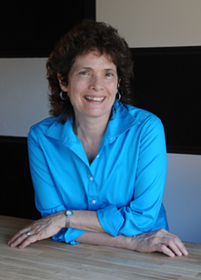 A woman in blue shirt sitting at table with arms crossed.